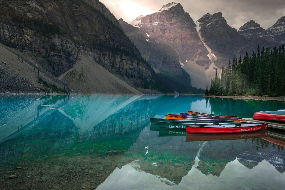 A row of colorful canoes docked on a pier next to a turquoise lake reflecting large, snow-capped mountains.