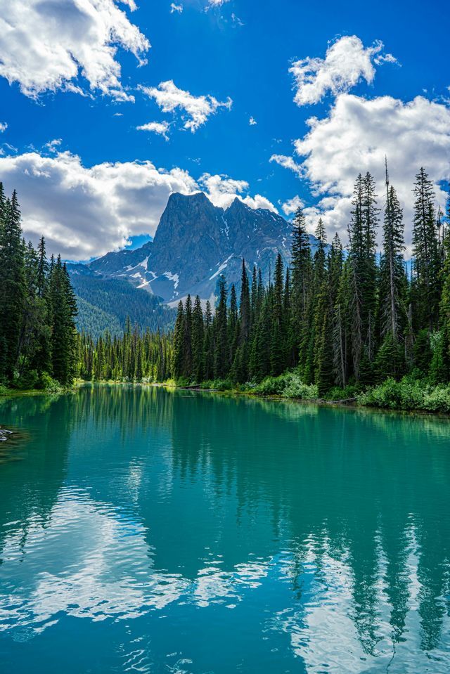 A calm turquoise lake reflects a pine forest and a jagged mountain peak with patches of snow under a blue, partly cloudy sky.