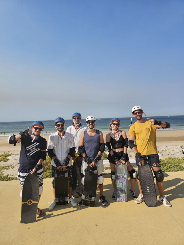 Un gruppo WeRoad in viaggio, con i caschi, posa con gli skateboard su un lungomare vicino a una spiaggia soleggiata.