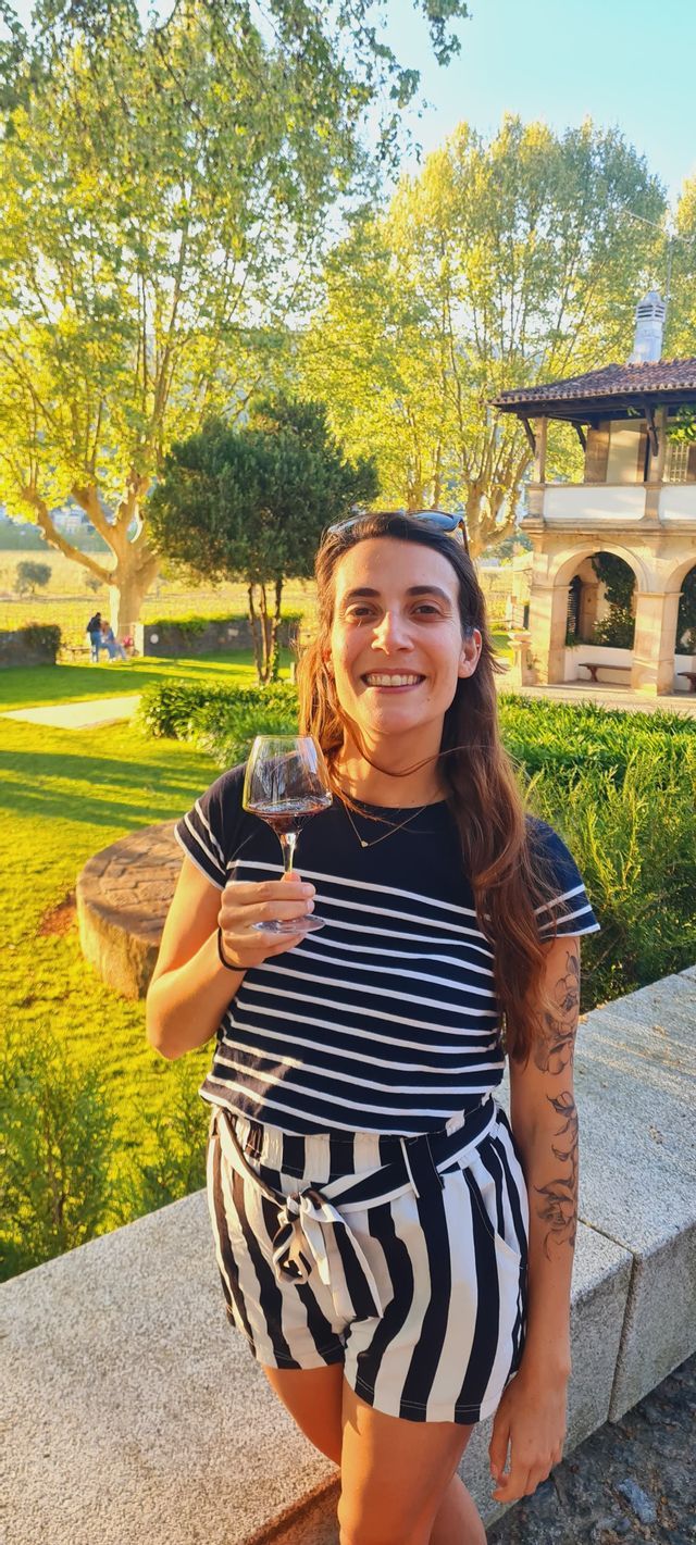 Una donna sorridente con un tatuaggio tiene in mano un bicchiere di vino rosso mentre si trova in un giardino soleggiato con alberi.