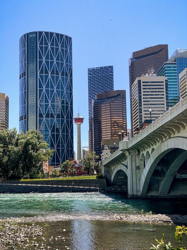 A stone arch bridge crosses a river with a modern city skyline featuring an observation tower in the background under a clear sky.
