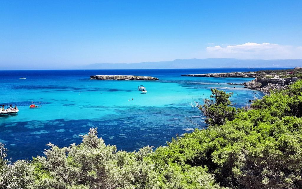 Veduta aerea di barche e nuotatori in una baia turchese circondata da una costa rocciosa e cespugli verdi sotto un cielo limpido.