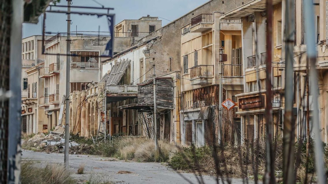 Una strada deserta fiancheggiata da edifici fatiscenti e abbandonati, con erbacce incolte e detriti a terra.