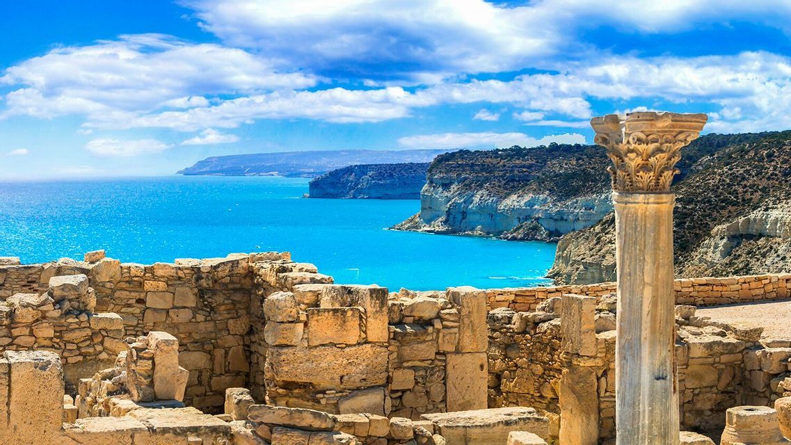 Antiche rovine in pietra e una colonna corinzia si affacciano su un mare blu brillante e scogliere sotto un cielo parzialmente nuvoloso.