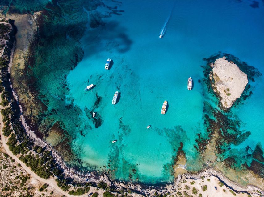 Una vista aerea di diverse barche ancorate in una baia cristallina e turchese vicino a una costa rocciosa e un piccolo isolotto.