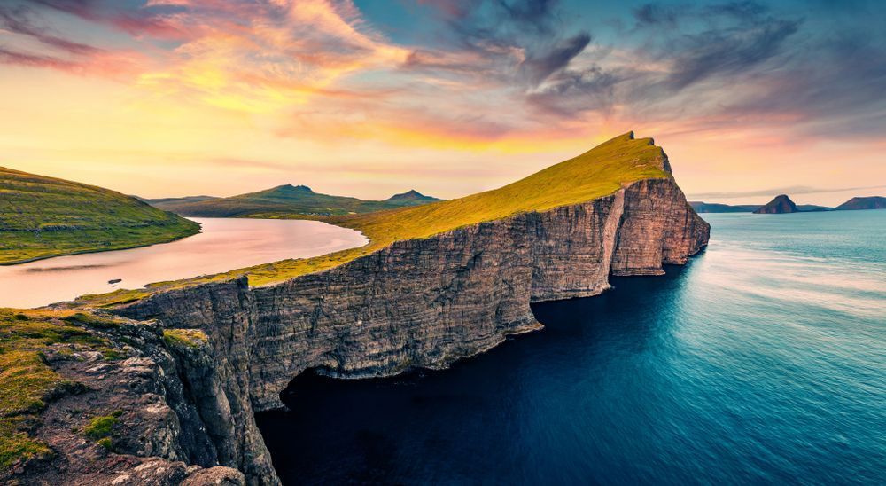 Un'alta scogliera verde separa un lago interno dall'oceano blu profondo sotto un cielo colorato al tramonto.