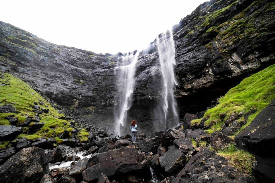 Una persona vista di spalle è in piedi su rocce scure alla base di una grande cascata che scende da una ripida scogliera muschiosa.