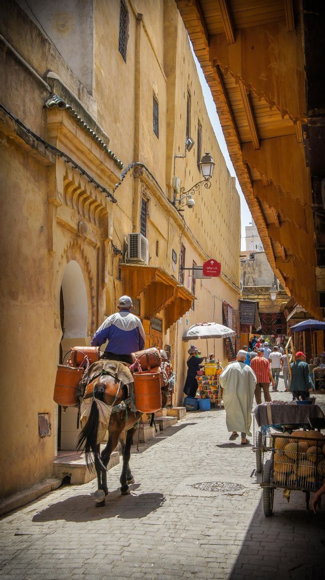 A man rides a donkey loaded with containers through a busy, narrow cobblestone alley flanked by yellow buildings.