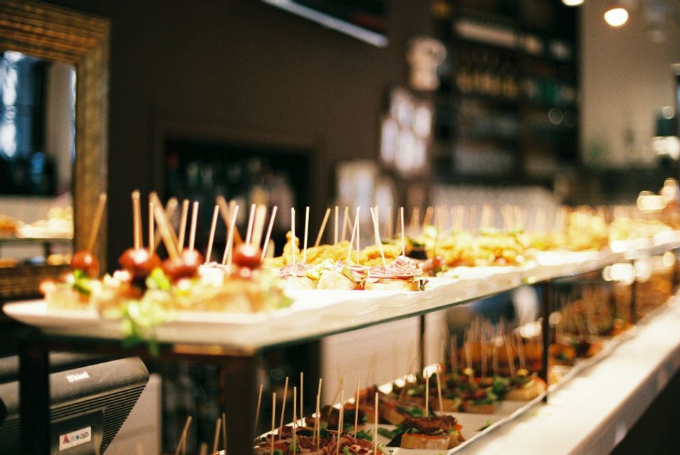 A variety of tapas with toothpicks arranged on long platters on a bar counter in a dimly lit restaurant.