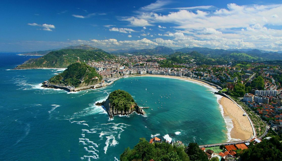 An aerial view of a coastal city along a crescent-shaped bay with a sandy beach, with a small island and green hills.