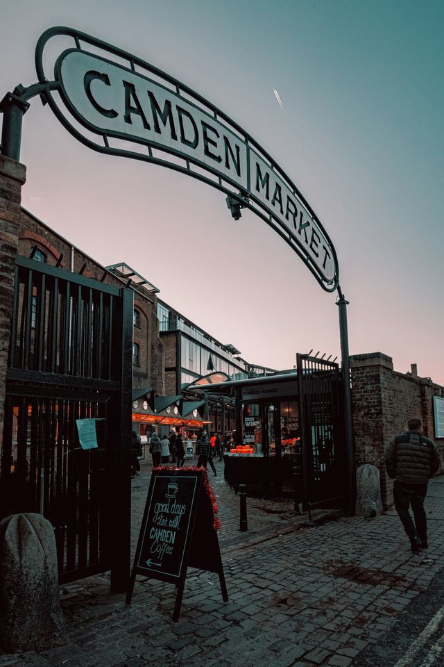 The entrance sign to Camden Market arches over a cobblestone street with people walking below at dusk.