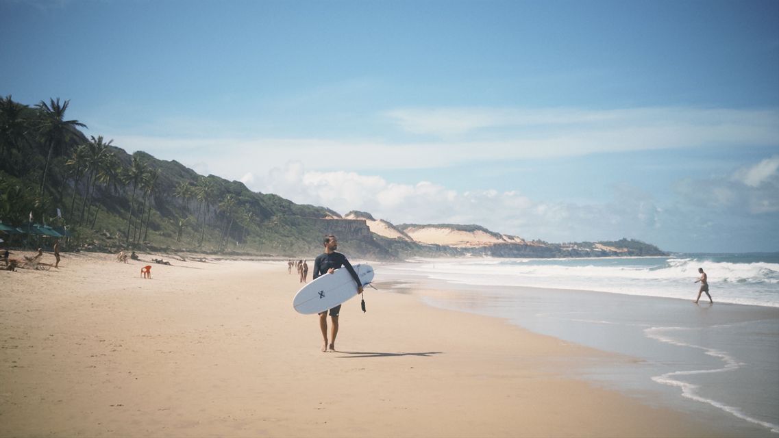 Un surfista in muta cammina su una spiaggia sabbiosa portando una tavola da surf, con onde e palme sullo sfondo.