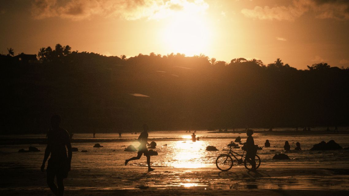 Sagome di persone su una spiaggia bagnata al tramonto, con una persona che corre e un'altra che cammina con una bicicletta in riva al mare.