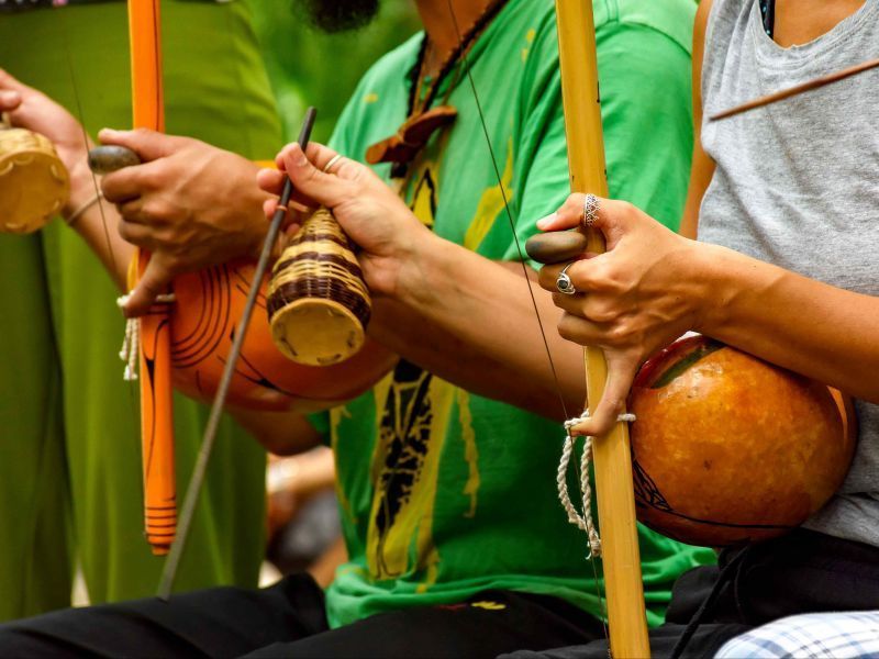 Un primo piano di due persone che suonano berimbau tradizionali e tengono in mano sonagli caxixi.
