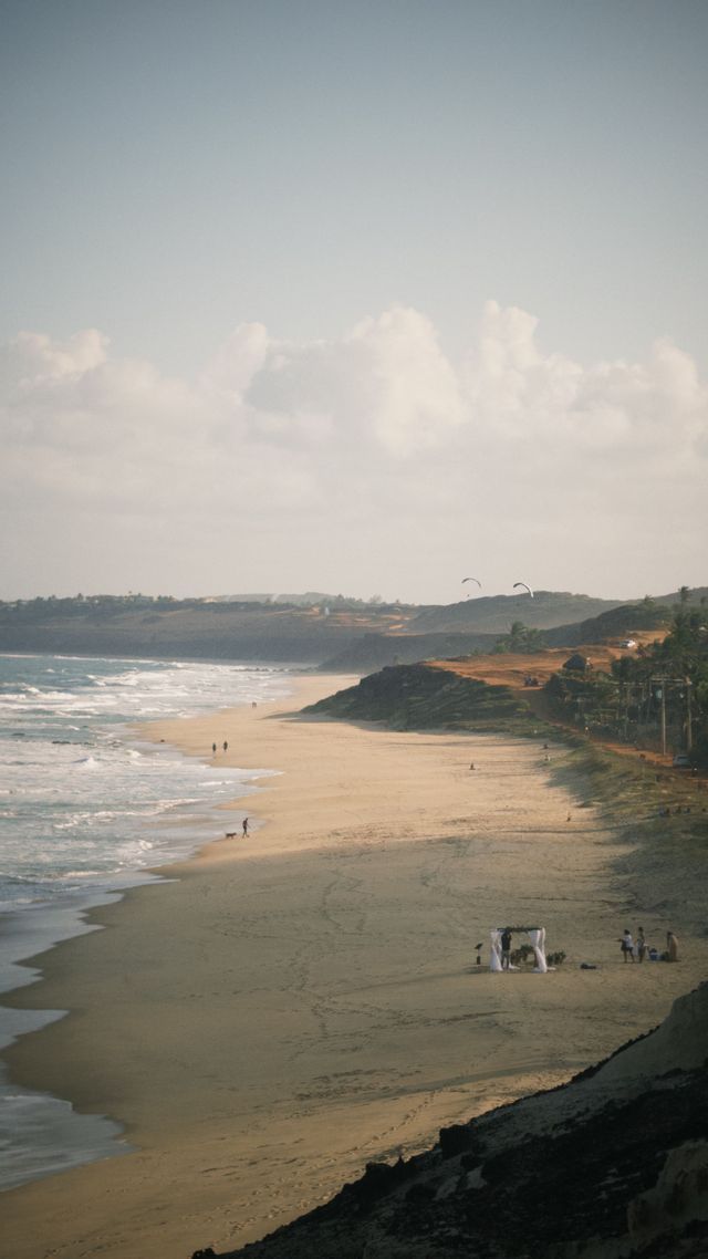 Una vista dall'alto di una lunga spiaggia sabbiosa con persone che passeggiano, un gazebo nuziale e kitesurfer in lontananza sotto un cielo nuvoloso.