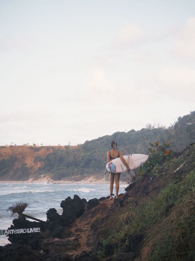 Una donna con una tavola da surf si erge su una scogliera rocciosa che domina una spiaggia con onde marine sotto un cielo nuvoloso.