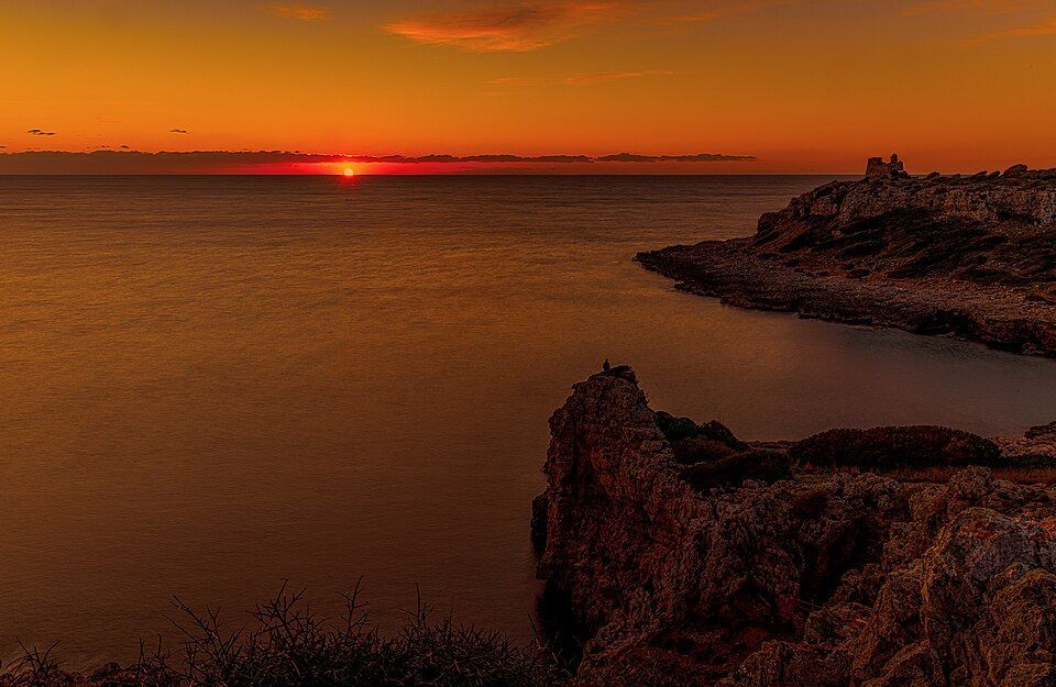 Una scogliera rocciosa con una piccola torre in pietra si staglia contro un tramonto arancione sul mare.