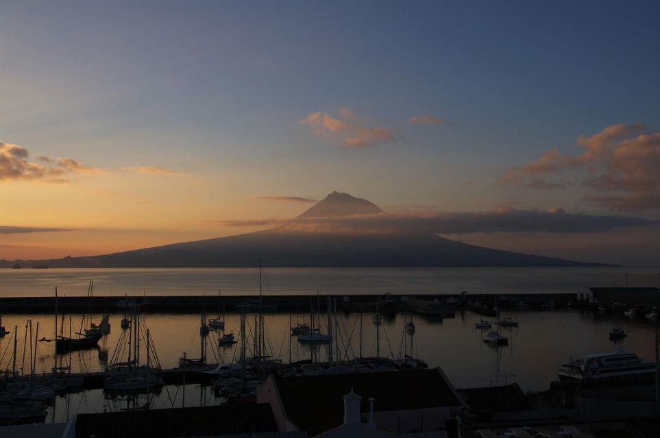 Un porto con barche a vela ormeggiate al tramonto, con un grande vulcano circondato da nuvole stagliato sull'acqua.