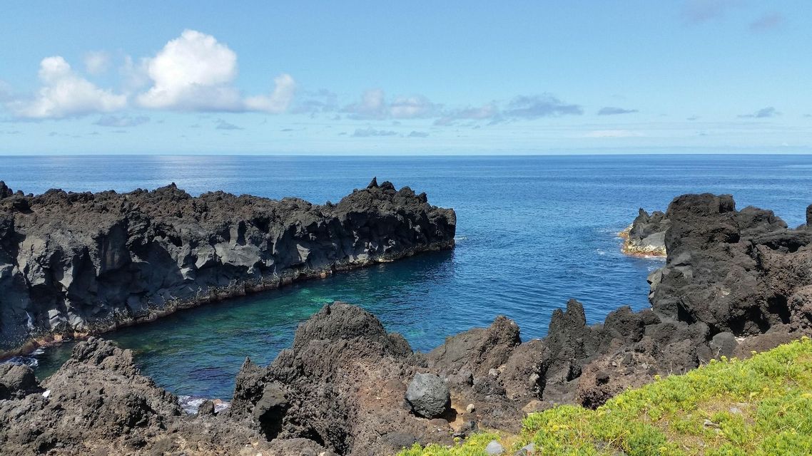 Rocce vulcaniche scure e frastagliate formano una piccola insenatura con acque limpide e turchesi in una giornata di sole lungo la costa.