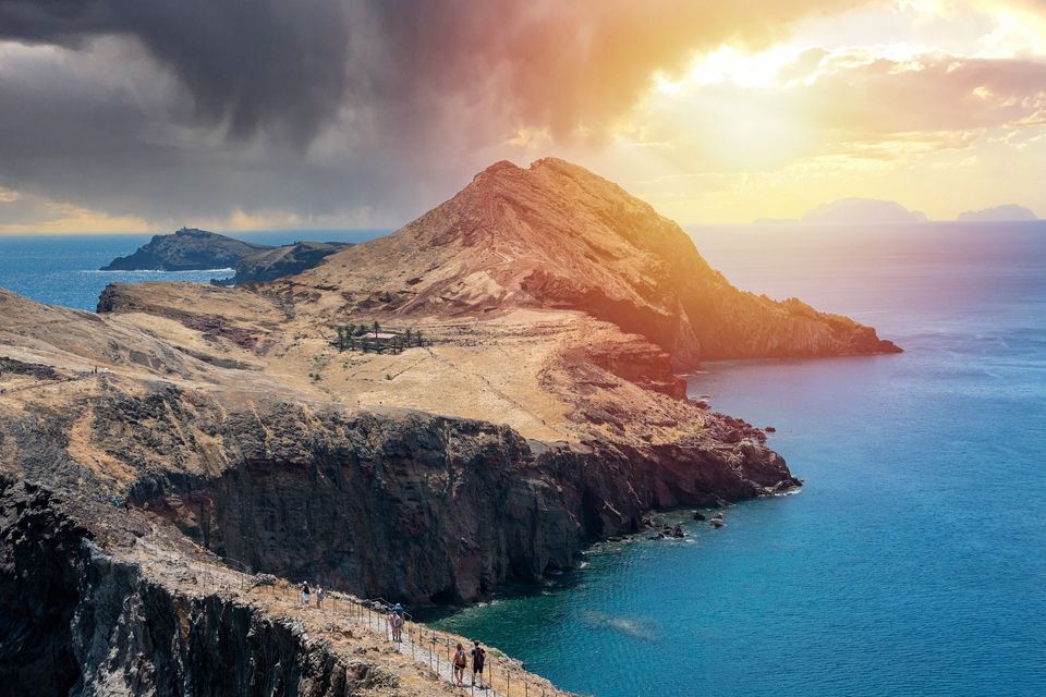 Un viaggio di gruppo WeRoad percorre uno stretto sentiero su una penisola rocciosa e costiera, sotto un cielo suggestivo con il sole che filtra tra le nuvole.