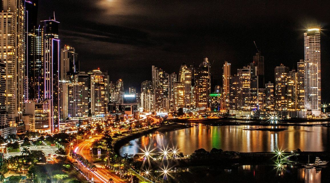 A city skyline of illuminated skyscrapers at night, with light trails on a road next to the water.