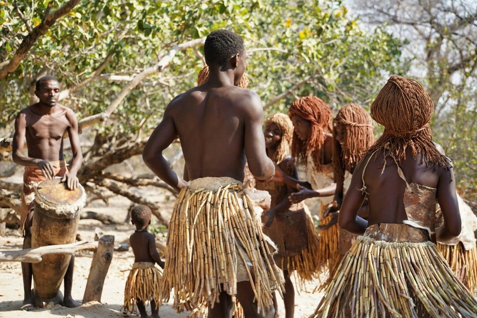 A group of tribal people in traditional attire, including reed skirts, dance while a man plays a drum outdoors.