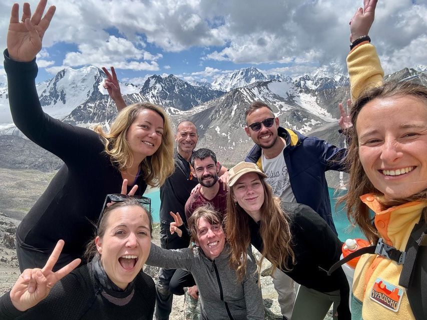 Un voyage de groupe WeRoad sourit pour un selfie avec un arrière-plan panoramique de montagnes enneigées et un lac turquoise.