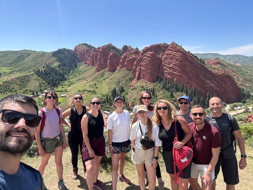 Un voyage de groupe WeRoad prend un selfie devant une formation rocheuse rouge et une vallée verdoyante et arborée sous un ciel bleu clair.