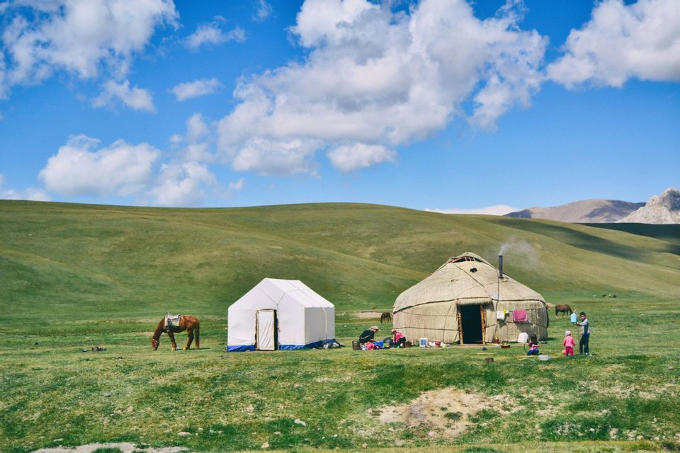 Une yourte traditionnelle et une tente blanche sont installées dans une vaste prairie verdoyante, avec des collines ondulantes en arrière-plan, et une famille ainsi qu'un cheval à proximité.