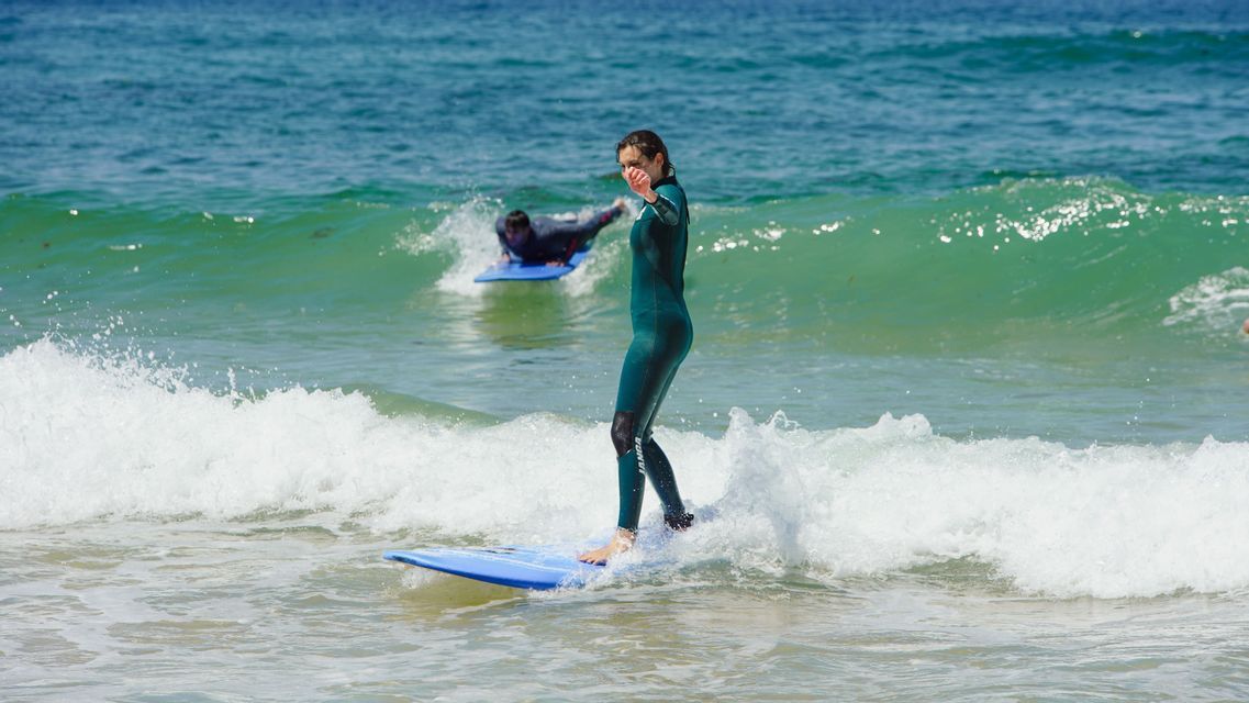 Una donna in muta verde si trova su una tavola da surf blu, cavalcando una piccola onda durante una giornata di sole in un viaggio di gruppo WeRoad.