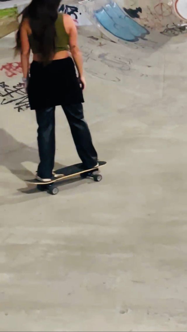 Una donna dai lunghi capelli scuri, vista di spalle, che fa skateboard in uno skatepark di cemento.