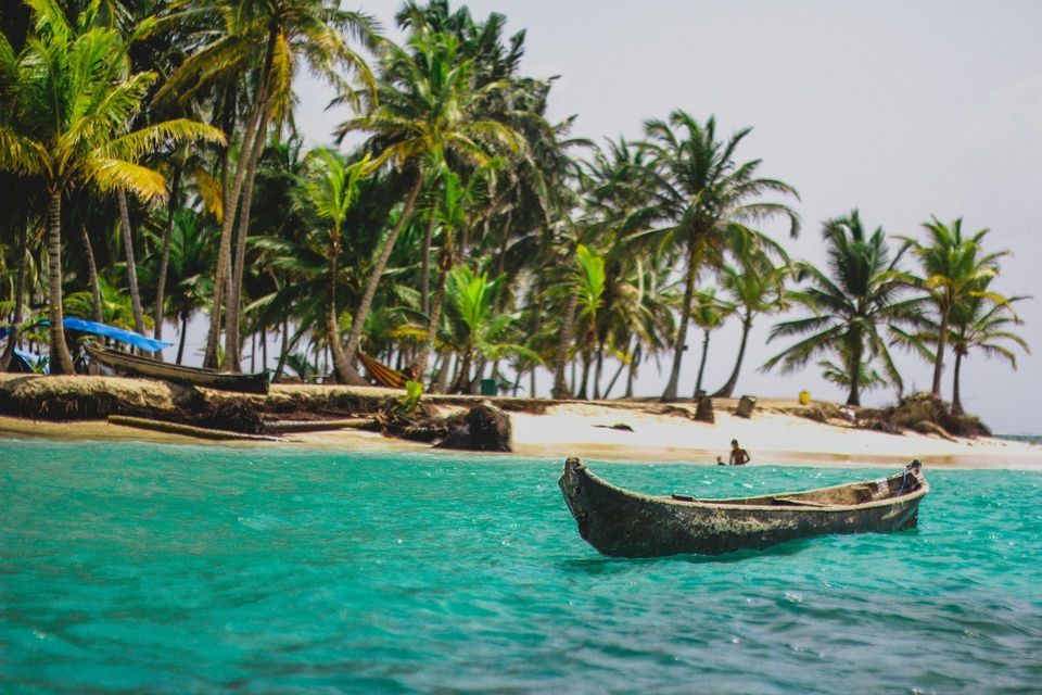 A wooden canoe floats on calm turquoise water in front of a sandy beach lined with tall palm trees.