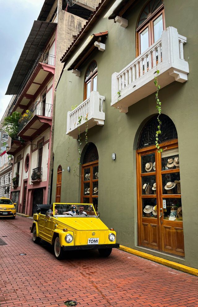 A yellow vintage convertible car drives on a red brick road next to colorful buildings with balconies.
