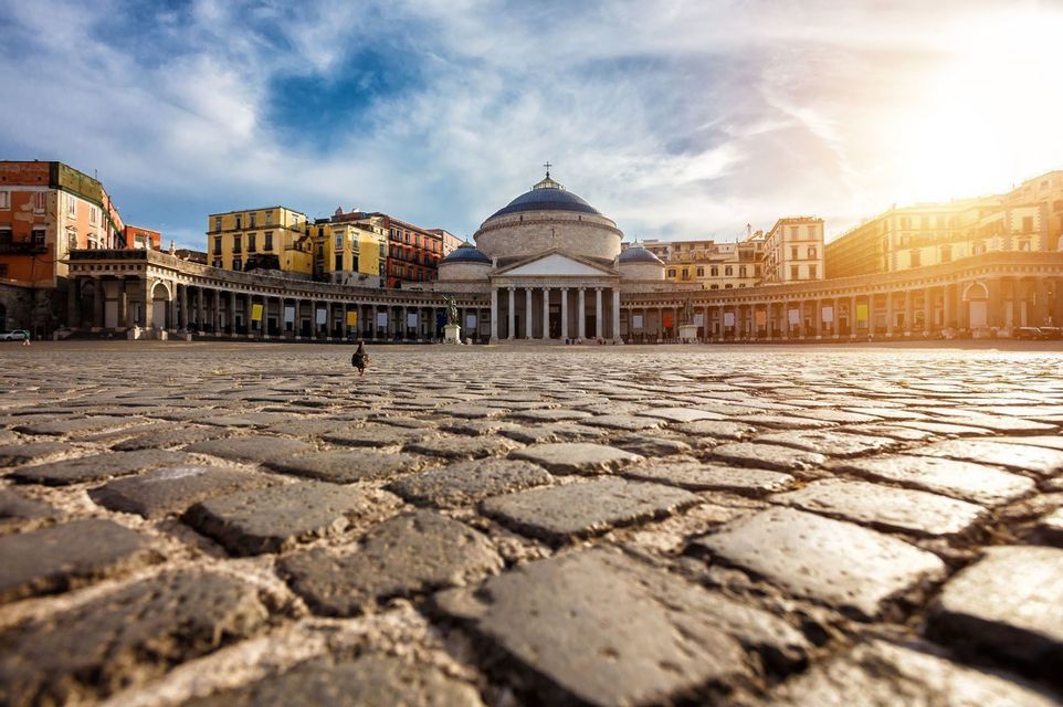 Una veduta dal basso di un piccione che cammina su una vasta piazza acciottolata, di fronte a un grande edificio a cupola con colonne.
