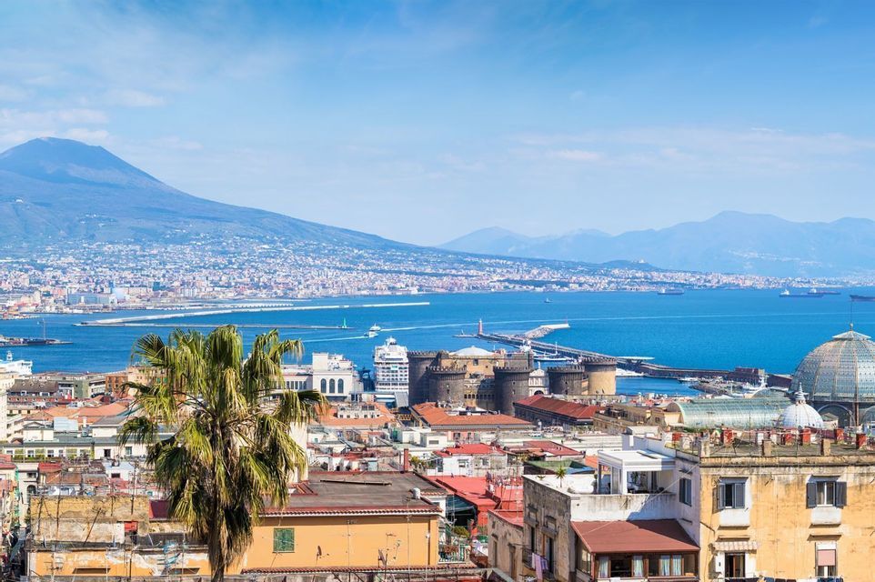 Vista dall'alto di una città costiera con un grande vulcano sullo sfondo, che mostra tetti, una palma e una baia blu brillante.