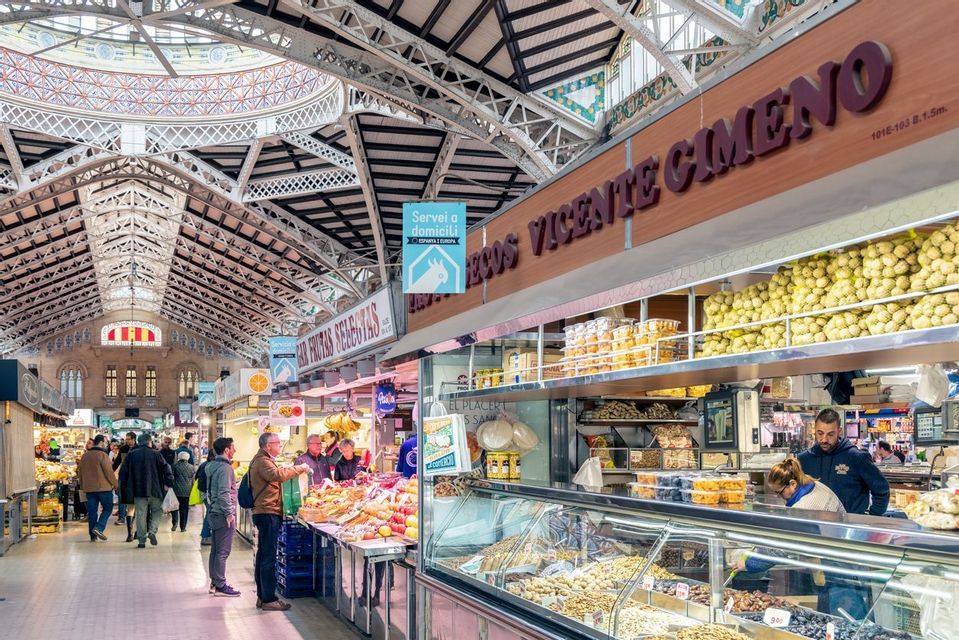 Des gens faisant des achats aux étals de nourriture dans une halle de marché intérieure animée, avec un haut plafond architectural orné.