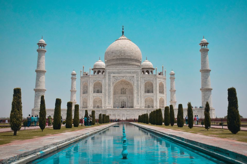 Il Taj Mahal di marmo bianco si riflette in una lunga vasca d'acqua, affiancato da giardini e alberi sotto un cielo azzurro e limpido.