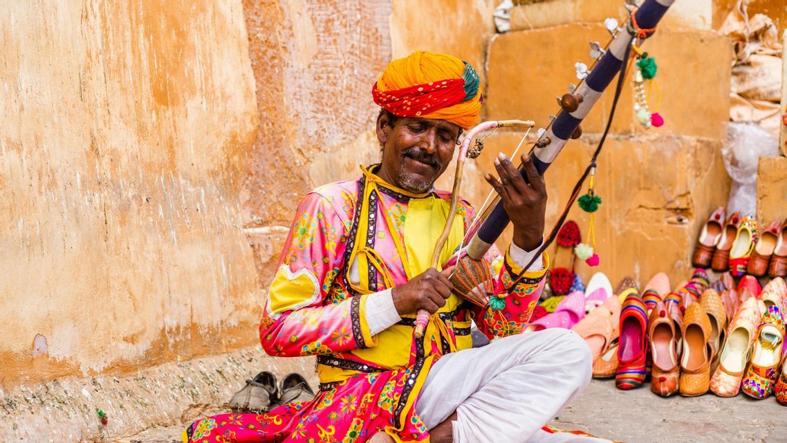 Un uomo con un turbante colorato e abiti tradizionali suona uno strumento a corde seduto accanto a un'esposizione di scarpe.