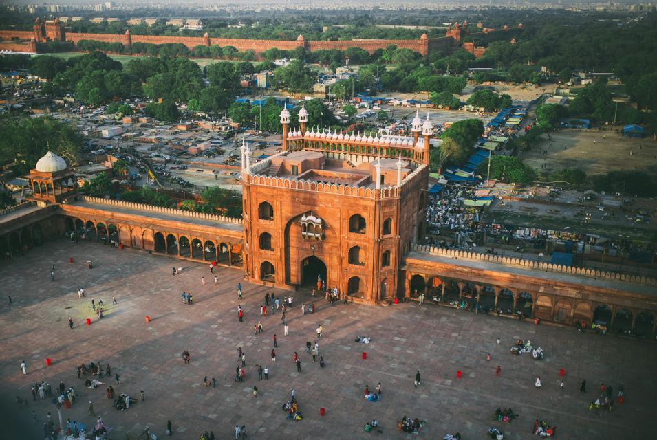 Veduta aerea di una moschea di arenaria rossa con persone nel suo ampio cortile, con una città e mura di un forte sullo sfondo.