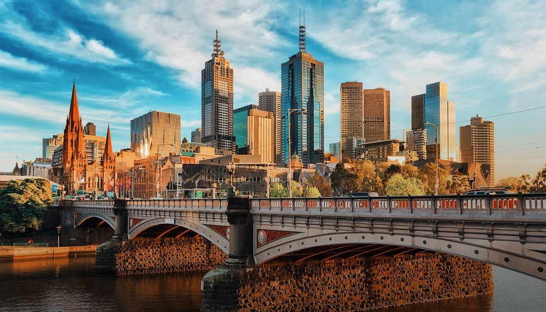 An ornate stone bridge crosses a river, with a city skyline featuring modern skyscrapers and a historic cathedral in the background.
