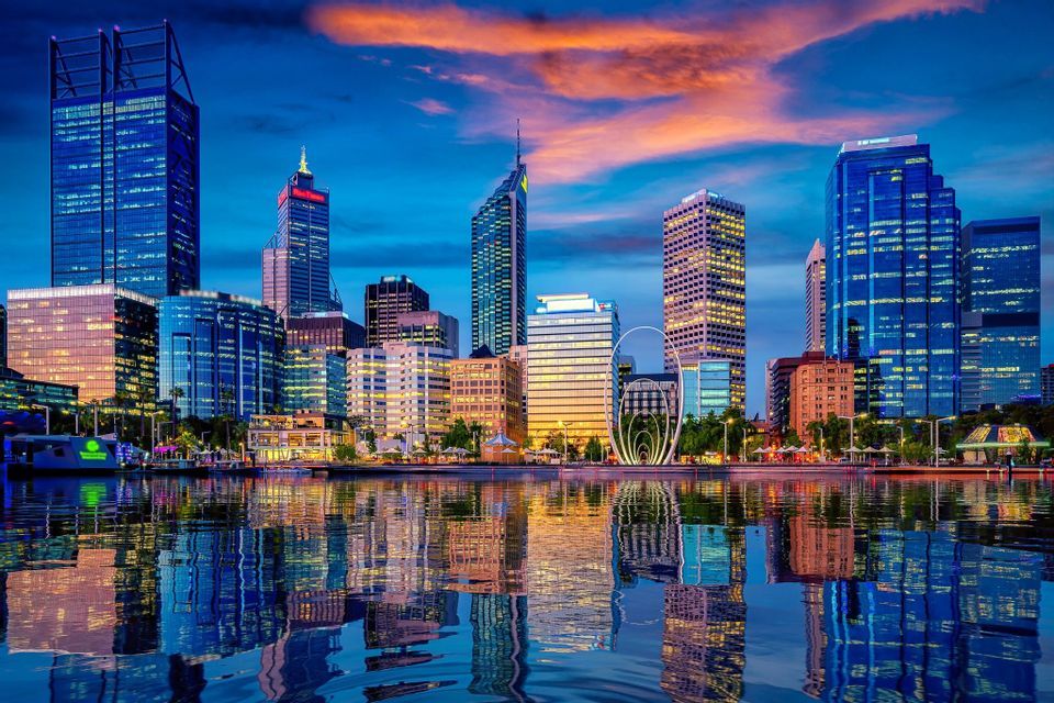 A modern city skyline with illuminated skyscrapers is reflected in the water below at dusk.