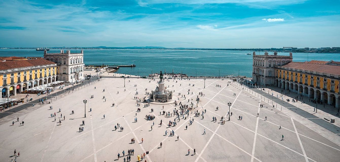 An aerial view of a large, sunlit plaza next to the sea, with people walking between historic buildings and a central monument.