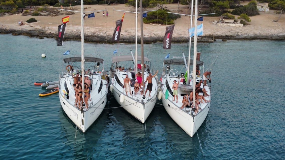 Un gruppo WeRoad in viaggio posa su tre barche a vela bianche ancorate fianco a fianco in una baia tranquilla vicino a una costa boscosa.