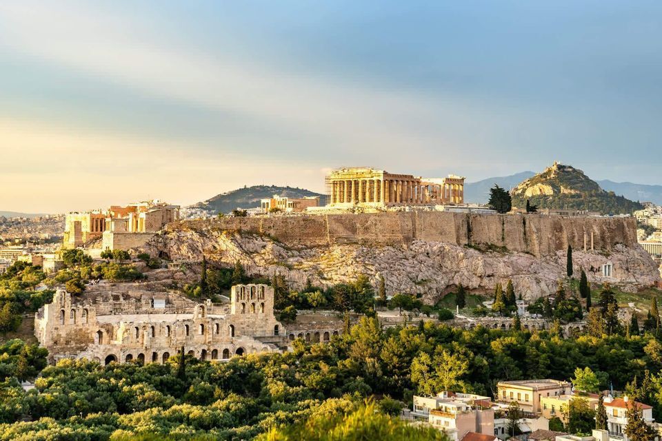 Antike griechische Ruinen auf einer felsigen Akropolis, von goldenem Licht beleuchtet, überblicken eine moderne Stadt und dichte grüne Bäume.