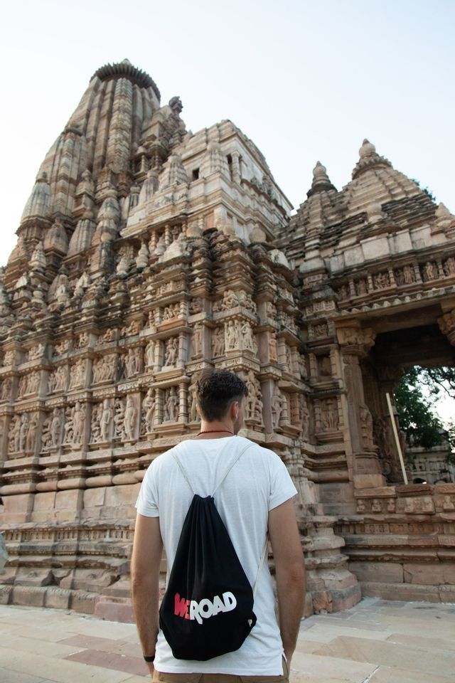 Un hombre visto de espaldas, vistiendo una camiseta blanca y una mochila WeRoad, de pie frente a un antiguo templo de piedra elaboradamente tallado.