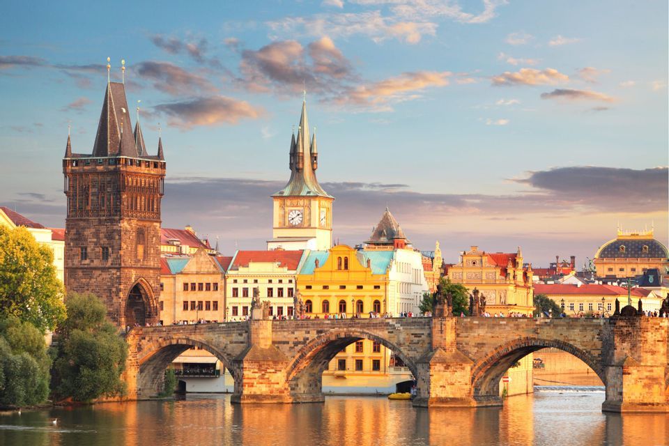 Un pont en arc de pierre avec des tours enjambe une rivière dans une ville historique, illuminé par la lumière chaude du coucher de soleil.