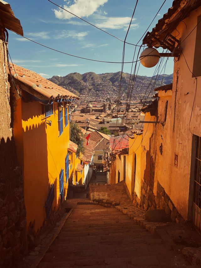 Un callejón de piedra estrecho y soleado, con escaleras, desciende entre edificios coloridos, revelando una vista de la ciudad y las montañas.