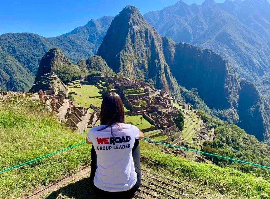 Una mujer con una camiseta de "WeRoad Group Leader" se sienta en una colina cubierta de hierba con vistas a ruinas antiguas anidadas entre escarpadas montañas verdes.