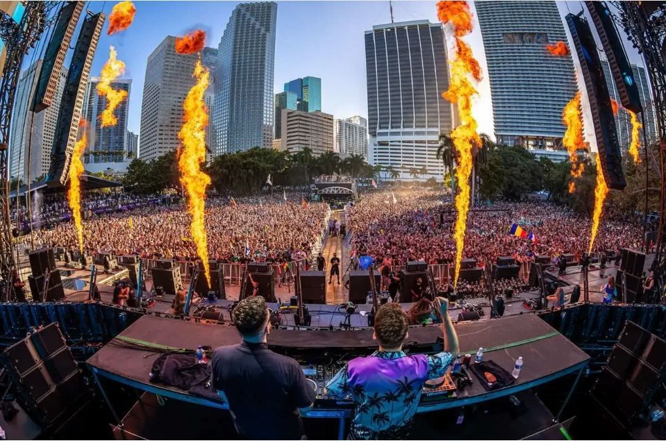 Due DJ si esibiscono su un palco per una folla immensa a un festival all'aperto con fuochi d'artificio che sparano fiamme, sullo sfondo dello skyline di una città.