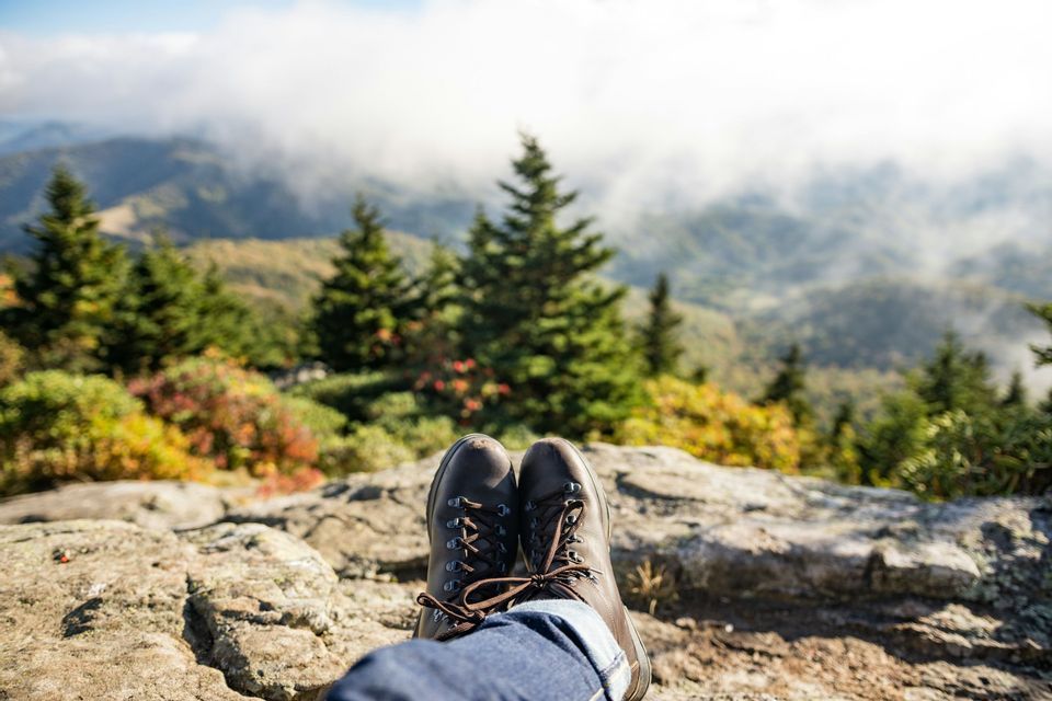 Le gambe di una persona con scarponi da trekking riposano su una sporgenza rocciosa, con vista su un paesaggio montuoso con alberi sempreverdi e nuvole basse.